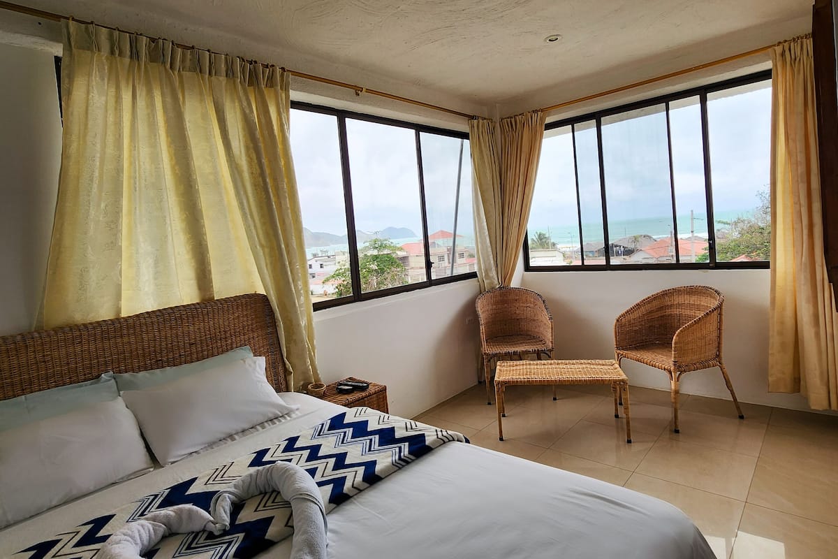 A hotel room with a large bed, two wicker chairs, a view of the ocean, and a balcony.