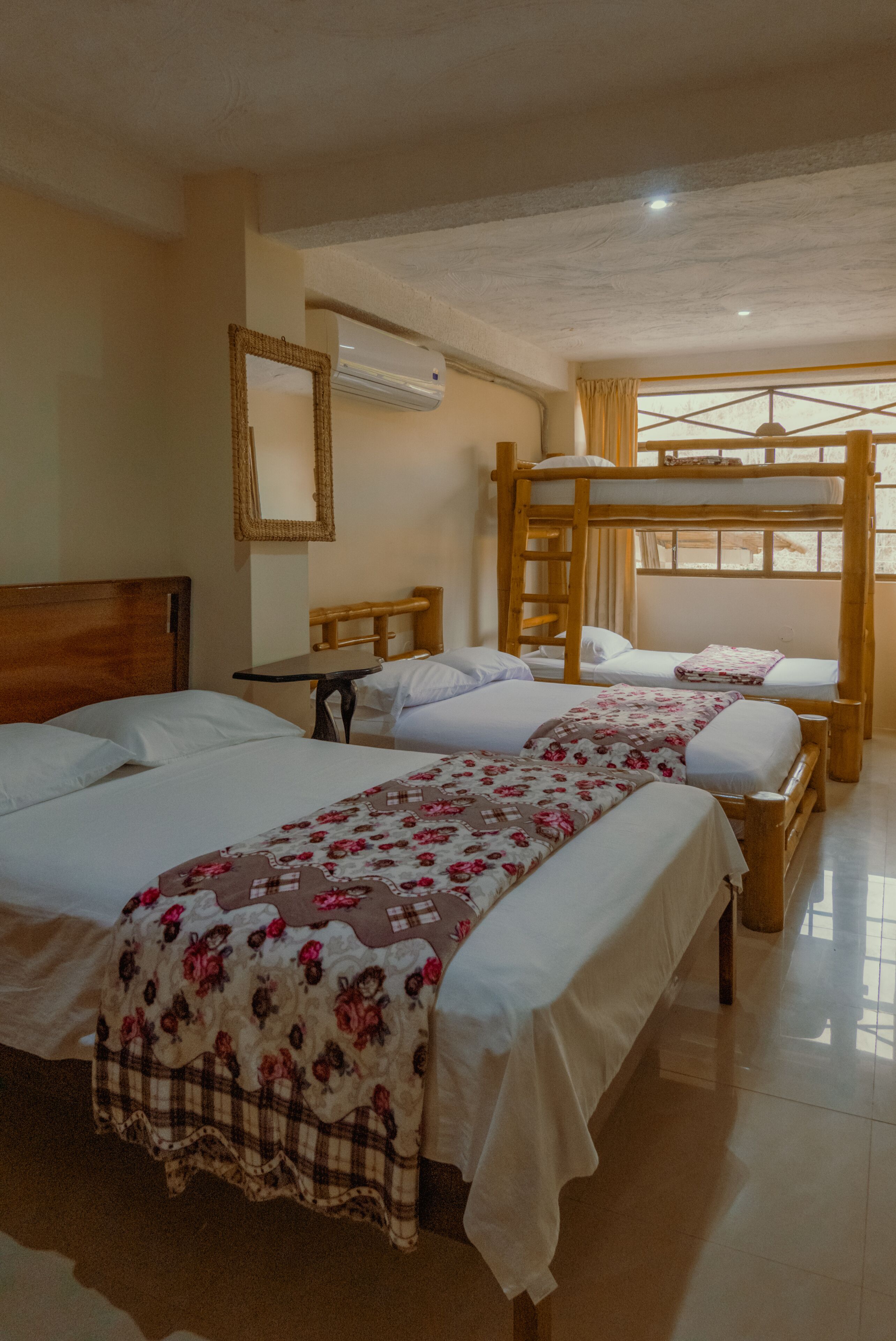 Habitación de hotel con tres camas, una litera de madera, una mesita, un espejo y una ventana con cortinas.
