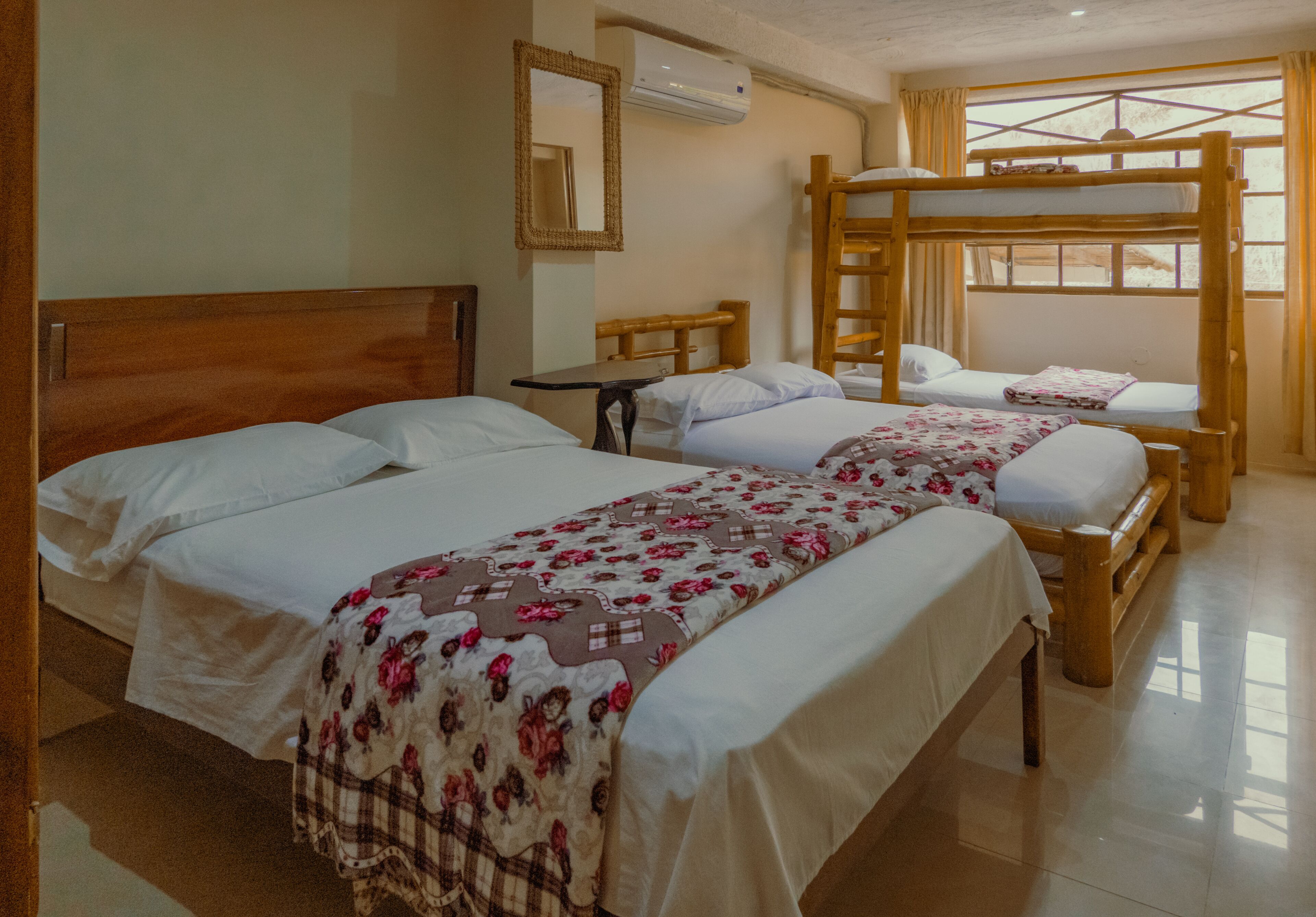 A hotel room with two single beds, a bunk bed, a wooden headboard, a small table, a mirror, and a window with curtains.