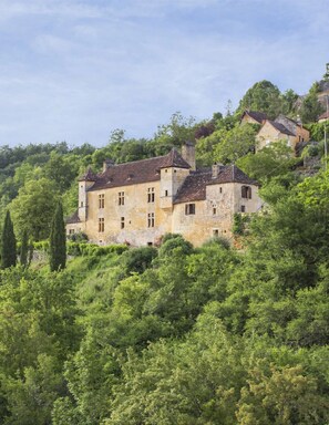 Un bâtiment historique doté de nombreuses fenêtres et d’une façade en pierre, entouré d’une végétation luxuriante et d’arbres.