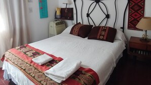 Un lit bien fait, recouvert d’une couvre-lit à motifs, un ventilateur de plafond, une lampe fixée au mur et une tapisserie rouge à motifs.
