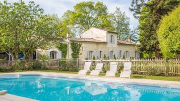 A pool with lounge chairs, a stone building with a tiled roof, and a wooden fence.