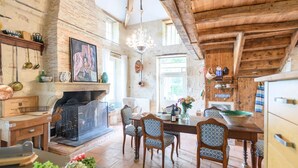 A rustic kitchen with exposed brick walls, wooden beams, a fireplace, and a dining area with patterned chairs. - Villa Du Fleuve - Four Bedroom Villa, Sleeps 8 (Flaujagues)