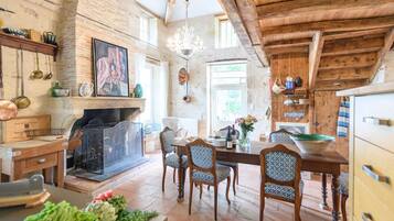 A rustic kitchen with exposed brick walls, wooden beams, a fireplace, and a dining area with patterned chairs.