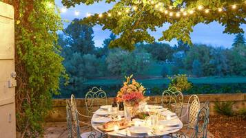 Un área de comedor al aire libre con una mesa preparada para dos, rodeada de sillas de metal, bajo un árbol con luces colgantes.