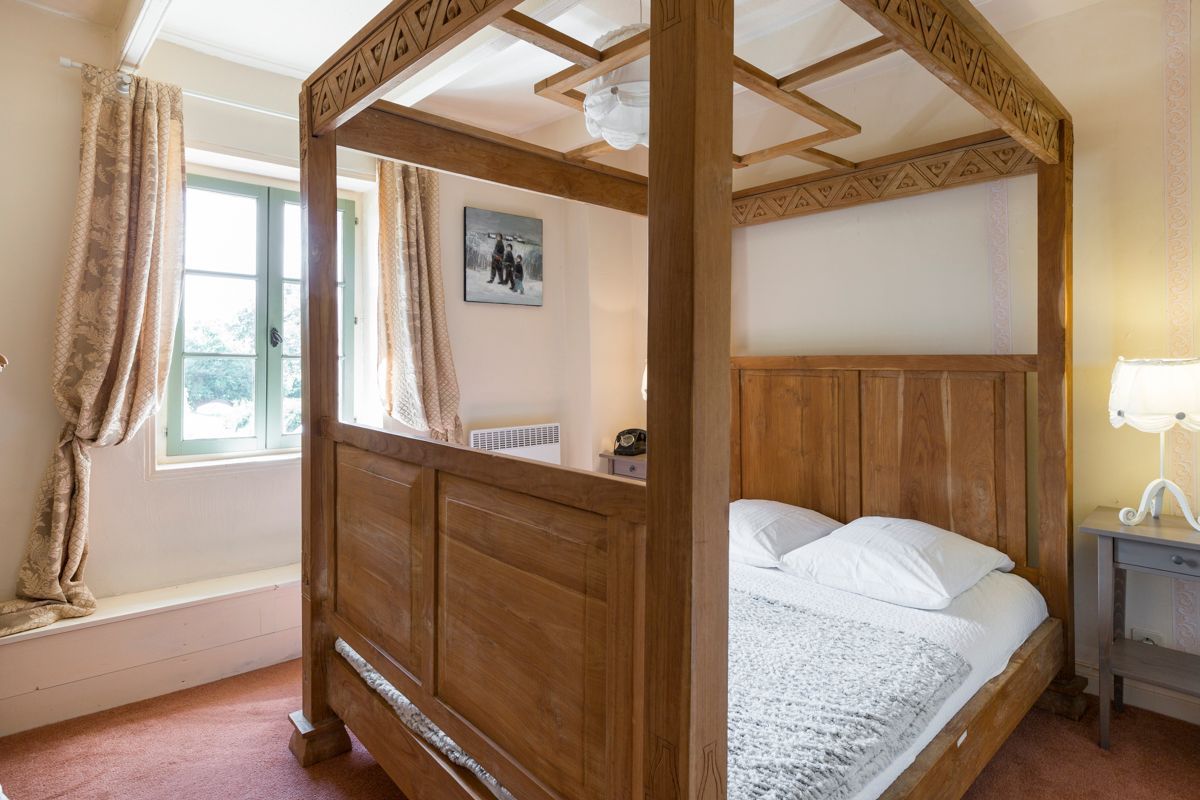 A four-poster bed with a wooden headboard, a bedside table with a lamp, and a window with curtains.