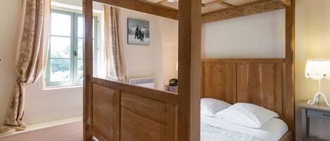 A four-poster bed with a wooden headboard, a bedside table with a lamp, and a window with curtains.