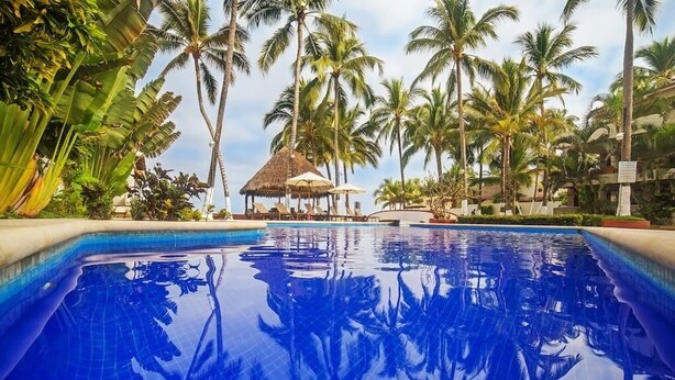 Ein klares, blaues Schwimmbecken, umgeben von Palmen und einem strohgedeckten Gebäude.