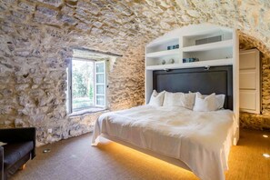 Quarto com parede de pedra, uma cama grande com roupa de cama branca, uma estante embutida branca e uma janela com cortinas.