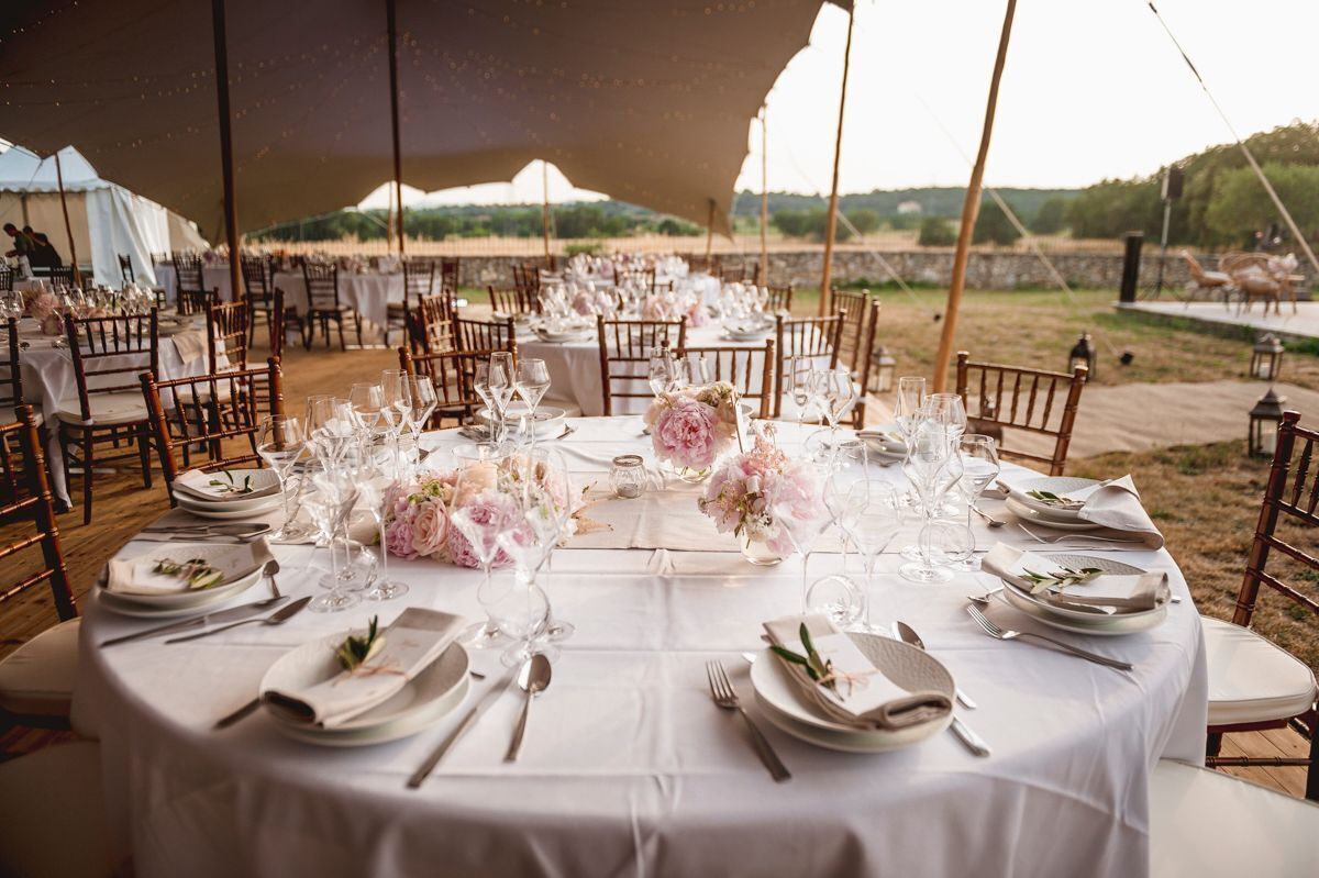 An outdoor dining area with round tables set for a formal event, surrounded by tents and chairs.
