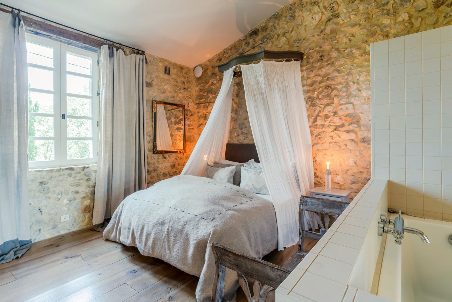 Ein Schlafzimmer mit Steinwand, einem Himmelbett, einem Nachttisch mit Lampe und Blick ins Freie durch ein Fenster.