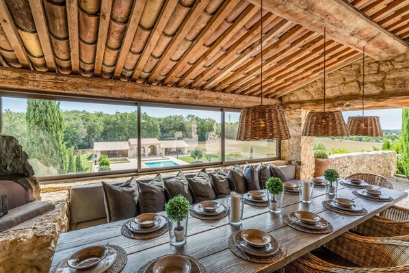 Un coin repas rustique avec une longue table dressée pour un repas, de grandes fenêtres donnant sur une piscine et un espace verdoyant, et un mur en pierre.