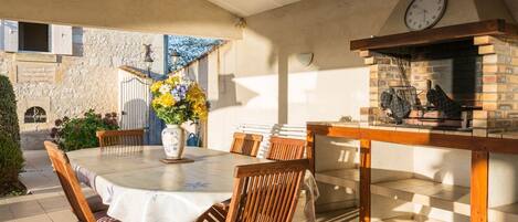 Un patio al aire libre con una mesa redonda, sillas de madera, una parrilla de ladrillo y un reloj en la pared.