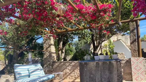 Un banco de madera con cojines a rayas, una mesa de piedra y una pérgola adornada con flores rosadas.