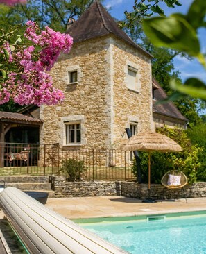 Un bâtiment en pierre avec une piscine, un parasol en chaume et des fleurs roses.