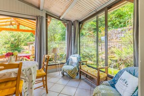 Een zonnige kamer met uitzicht op een tuin, een eettafel met stoelen en een gezellige zithoek met kussens.