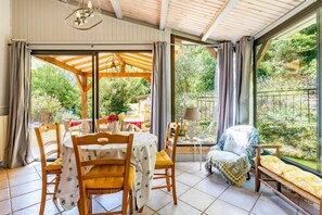 Une pièce baignée de lumière naturelle, avec une table en bois, des chaises et une vue sur un jardin, visible à travers de grandes fenêtres.