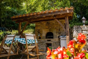 Een buitenkeuken van natuursteen met een houten pergola erboven, een tafel met geruit tafelkleed en een rustieke omgeving.