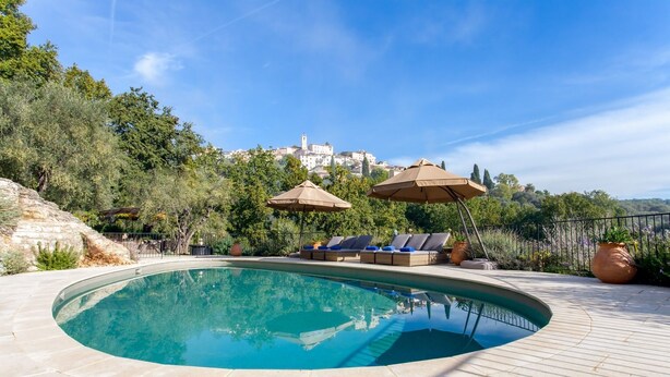 Una piscina con sillas de descanso y sombrillas, rodeada de vegetación y con vistas a edificios al fondo.