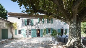 Un bâtiment blanc aux volets verts, une cour recouverte de gravier et un grand arbre qui offre de l’ombre.