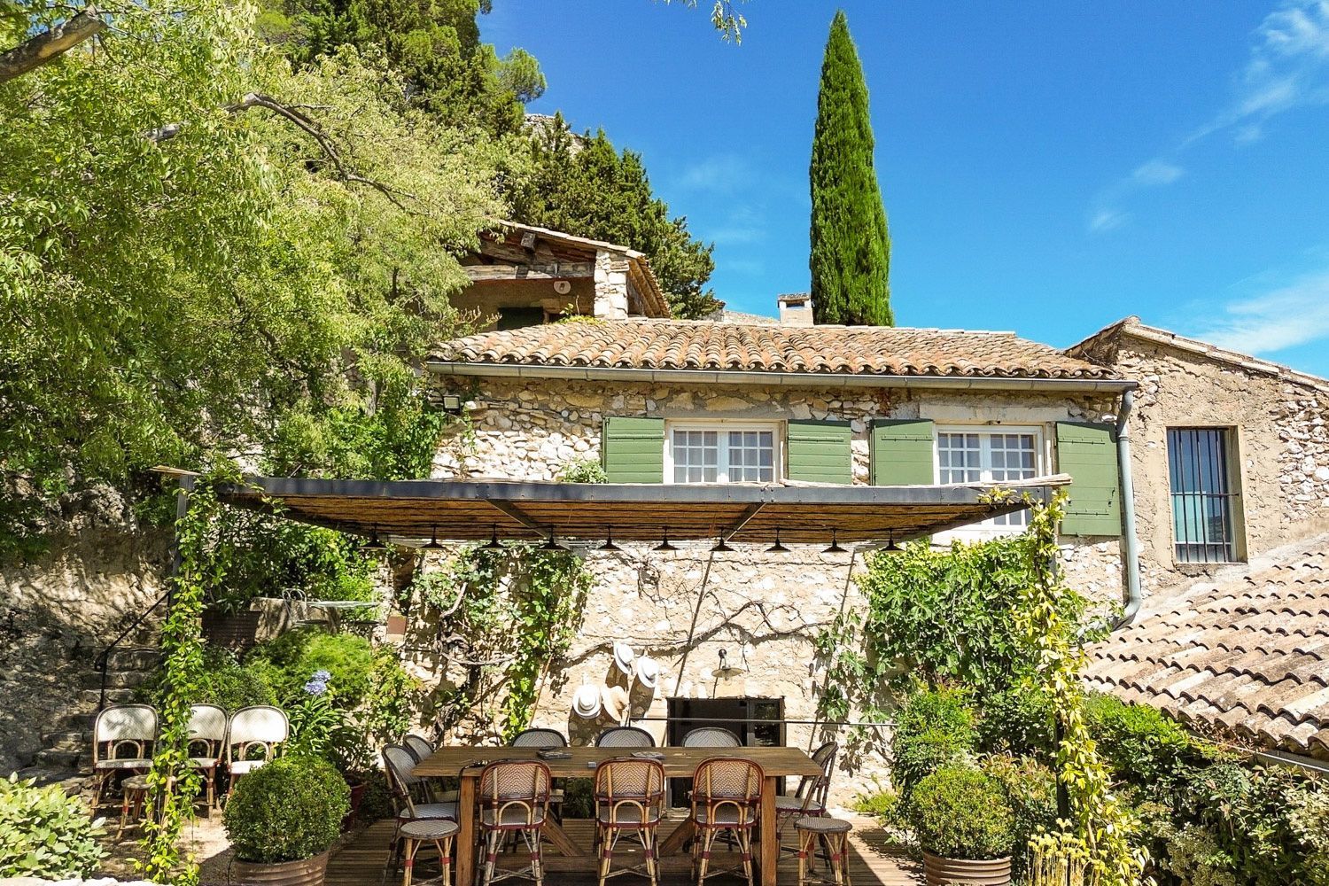 A rustic stone house with a tiled roof, a wooden pergola, and a dining area with wicker chairs.