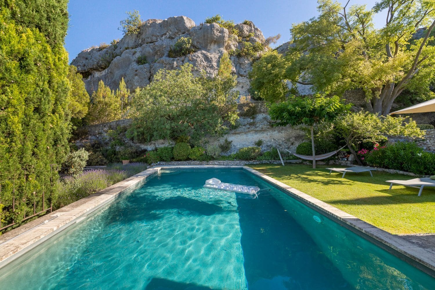 A rectangular swimming pool with clear blue water, surrounded by lush greenery and a rocky cliff in the background.