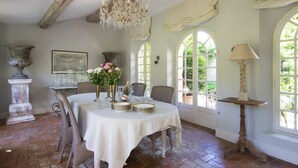Une salle à manger avec une table ronde dressée pour quatre personnes, entourée de chaises en osier. Un lustre est suspendu au-dessus de la table, et la pièce est dotée de grandes fenêtres avec des rideaux.
