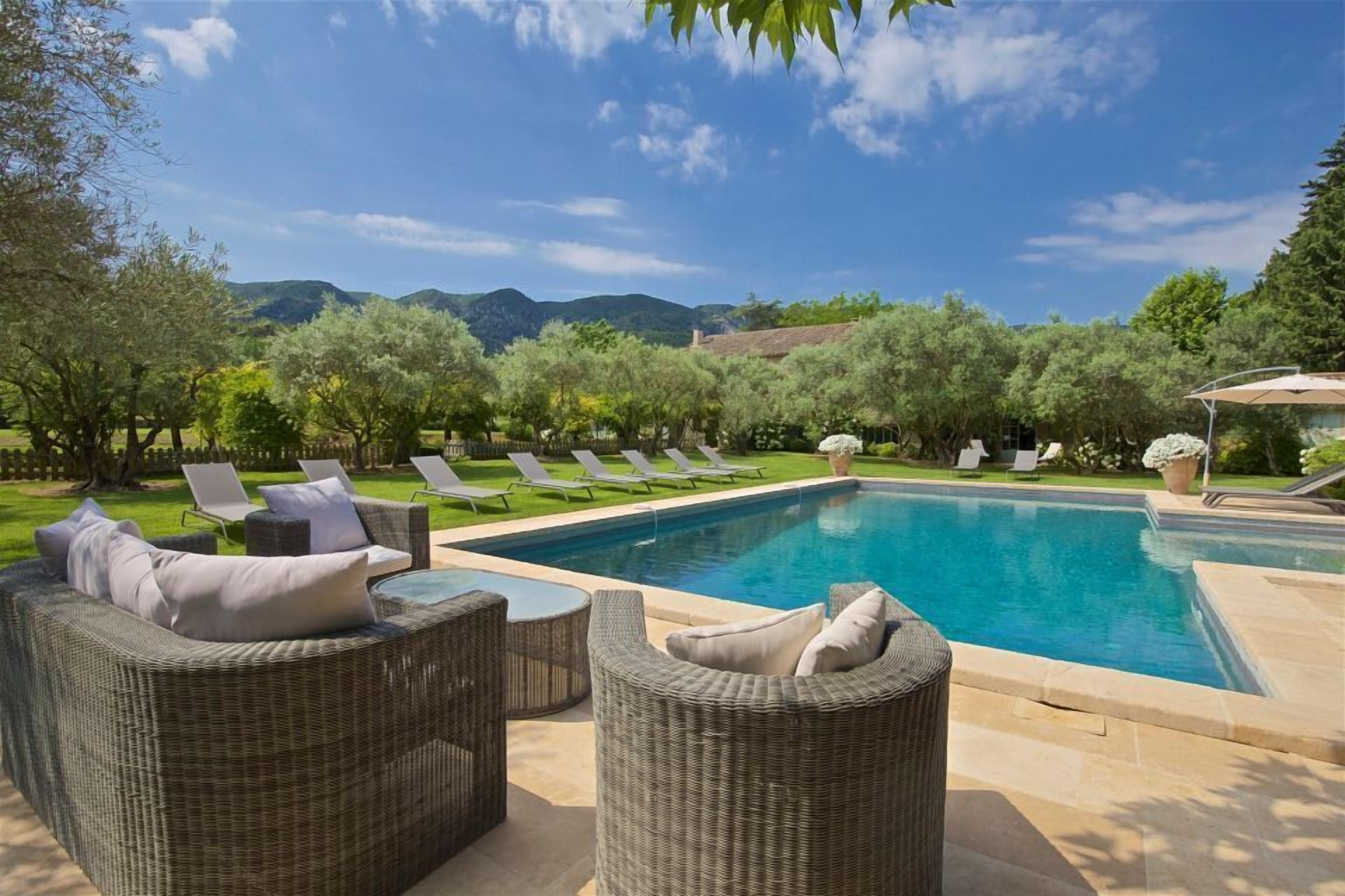 A pool area with wicker furniture and lounge chairs, surrounded by trees and hills in the background.
