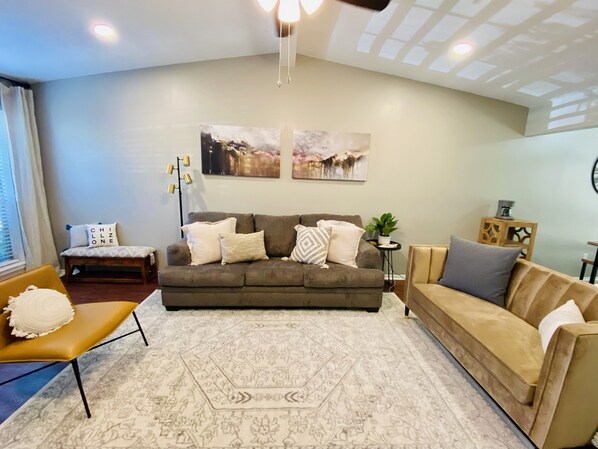 A living room with a sofa, a coffee table, and a patterned rug. There are two wall-mounted artworks and a floor lamp. - *New* Welcome to the Cheerful 2 Bedroom Duplex (Tyler)