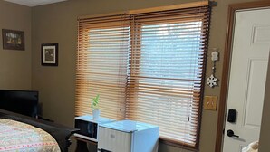 A bedroom with a bed, a small refrigerator, a desk with a computer, and a window with blinds.