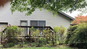 A house with a wooden deck, surrounded by greenery and a lawn.