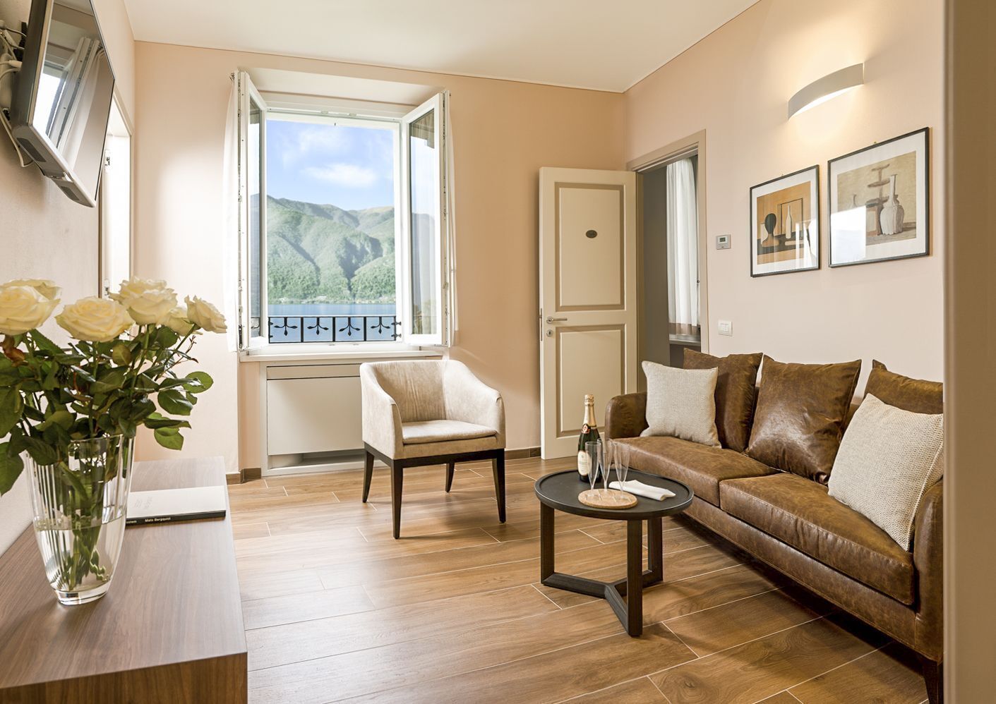 A living room with a wooden floor, a brown leather sofa, a white armchair, a small round table with a bottle and glasses, and a window with a view of green hills.