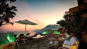 A rooftop terrace with lounge chairs, a glass table, and a view of the sunset over the ocean.