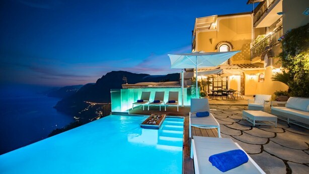A poolside area with lounge chairs and a table, overlooking a scenic coastline at dusk.