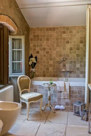 Un baño con bañera, una silla, una mesita con un florero y una zona de ducha.