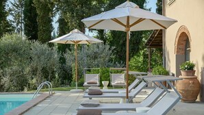 Zona de piscina con sillones de descanso y sombrillas, piscina y pared de ladrillo con un ventanal arqueado.