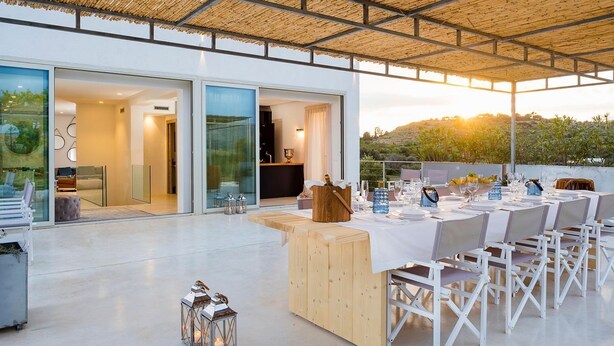 An outdoor dining area with a long table set for a meal, a wooden bar counter, and a view of the landscape.