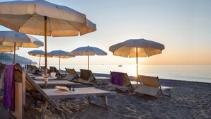 Un área de descanso frente a la playa con múltiples tumbonas y sombrillas, una mesa con una taza y un plato, y vistas al océano al atardecer.