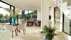 A modern living room with a glass staircase, a dining area with white chairs, and a large window offering a view of greenery. - Villa Incanto - Four Bedroom Villa, Sleeps 8 (Plemmirio)