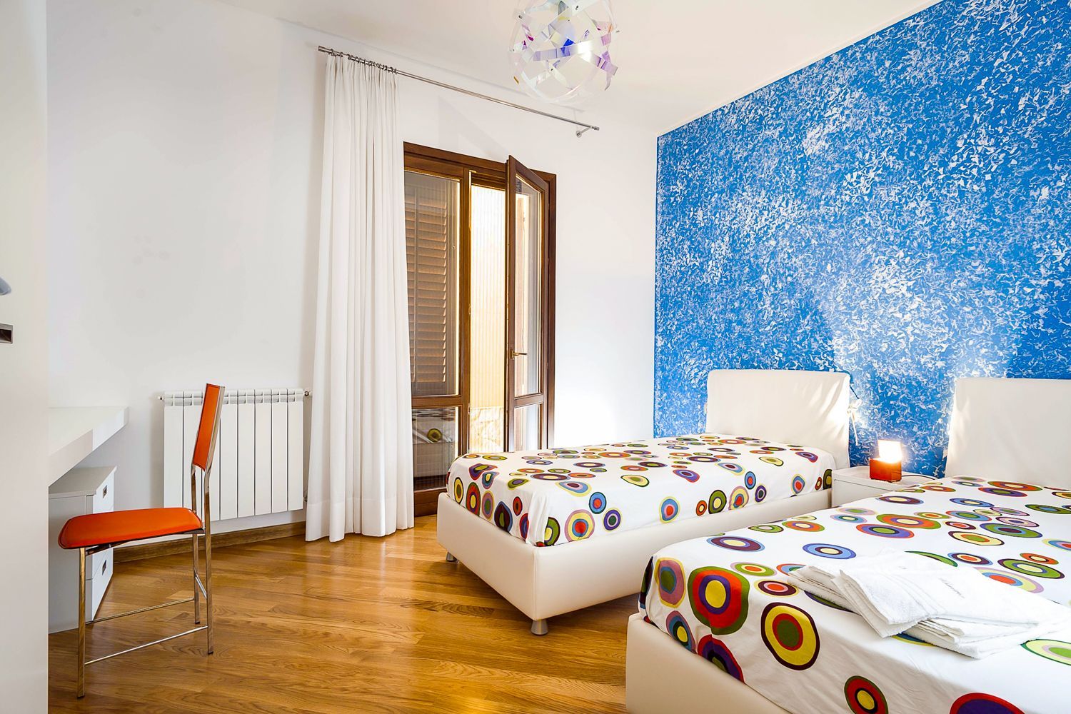 Un dormitorio con dos camas, una silla, un radiador, una ventana con cortinas y una pared azul con textura.