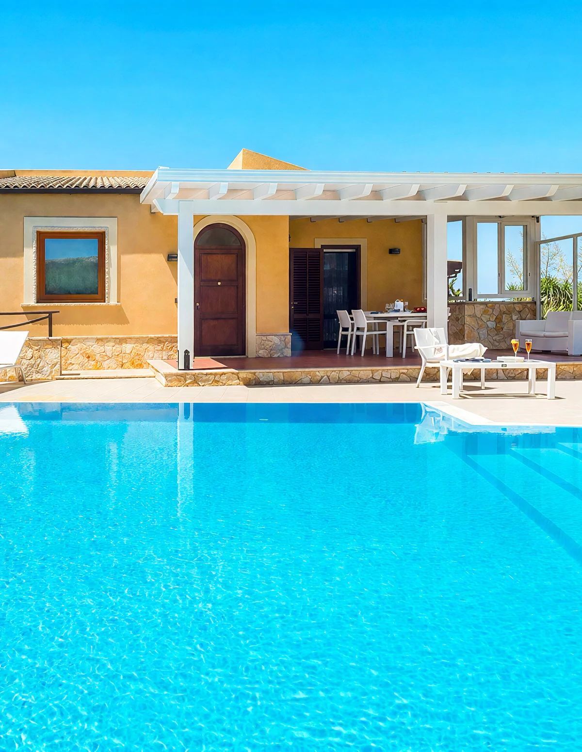 Ein Poolbereich mit einem klaren blauen Pool, einer überdachten Terrasse mit Sitzgelegenheiten und einer Steinmauer.