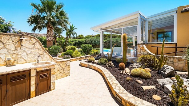 Een moderne buitenkeuken met stenen aanrechtbladen en een ingebouwde barbecue, omgeven door een aangelegde tuin met diverse cactussen en struiken.