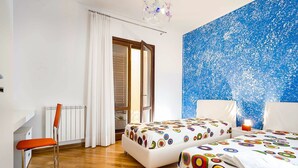 Een slaapkamer met twee bedden, een stoel, een radiator, een raam met gordijnen, en een blauwe wand met structuur.