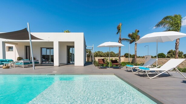 Ein moderner Poolbereich mit klarem Wasser, Liegestühlen, Sonnenschirmen und einem Gebäude mit großen Fenstern.