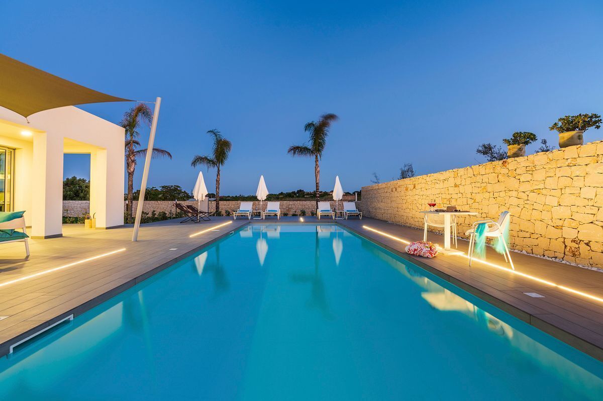 Una piscina amb gandules i ombrelles, envoltada per una paret de pedra i palmeres.