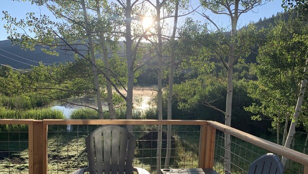 A wooden deck with two Adirondack chairs and a small table, overlooking a natural landscape with trees and a body of water.