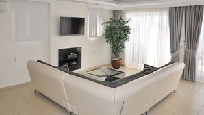 A modern living room with a white sectional sofa, a flat-screen TV mounted on the wall, a fireplace, and a large potted plant. - Villa Gunes - Three Bedroom Villa, Sleeps 6 (Ölüdeniz Belediyesi)