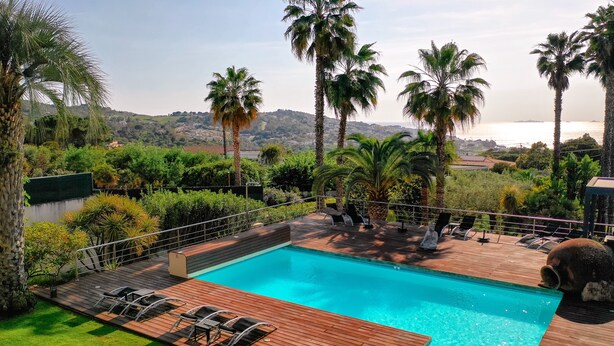 A swimming pool with a wooden deck, surrounded by palm trees and a view of the sea. - Villa 6pers splendid sea view and magnificent landscaped park in the peace with swimming pool (Grosseto-Prugna)