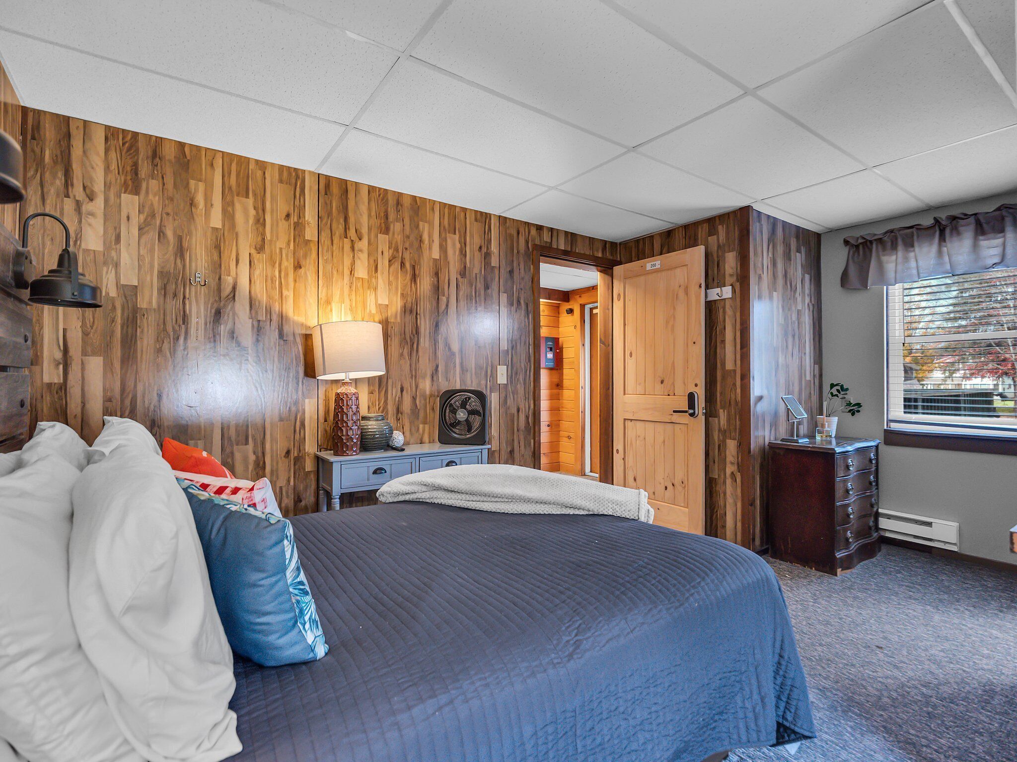 Une chambre avec un grand lit, des murs recouverts de lambris en bois, une table de chevet avec une lampe, une commode et une fenêtre avec des rideaux.