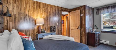 Une chambre avec un grand lit, des murs recouverts de lambris en bois, une table de chevet avec une lampe, une commode et une fenêtre avec des rideaux.